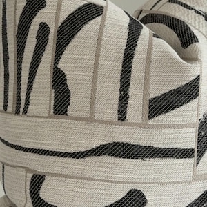 May include: Decorative throw pillow with a neutral beige background and a bold black abstract pattern. The design features thick, curved lines and rectangular shapes, creating a modern, geometric aesthetic. The pillow is made of a textured fabric.