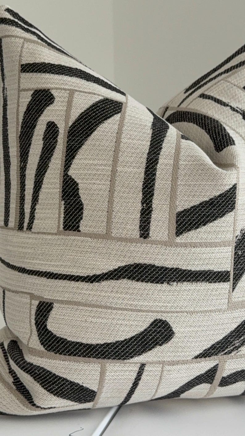 May include: Decorative throw pillow with a neutral beige background and a bold black abstract pattern. The design features thick, curved lines and rectangular shapes, creating a modern, geometric aesthetic. The pillow is made of a textured fabric.