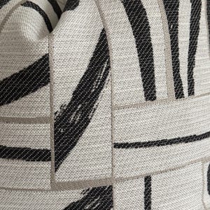 May include: Close-up of a textured pillow with a geometric pattern. The design features thick black brushstrokes and thin gray lines on a light gray background. The fabric has a woven appearance, creating a modern aesthetic.
