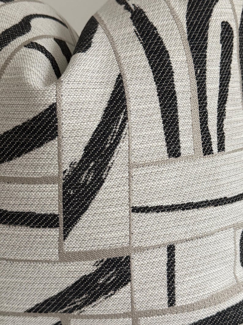 May include: Close-up of a textured pillow with a geometric pattern. The design features thick black brushstrokes and thin gray lines on a light gray background. The fabric has a woven appearance, creating a modern aesthetic.