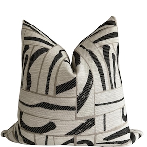 May include: A decorative throw pillow with a geometric pattern. The pillow features a beige background with black and gray abstract brushstroke designs. The pillow is square and has a soft, textured appearance.