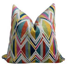 May include: A square throw pillow featuring a vibrant geometric pattern of interlocking chevrons and diamonds. The design incorporates a multi-colored palette of teal, mustard yellow, burnt orange, magenta, navy blue, and white, with a soft velvet texture.