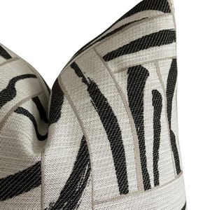 May include: Close-up of a decorative pillow with a geometric pattern. The pillow features a light beige background with bold black brushstroke-style stripes and thin, light gray lines. The pillow has a textured appearance and a soft, inviting look.