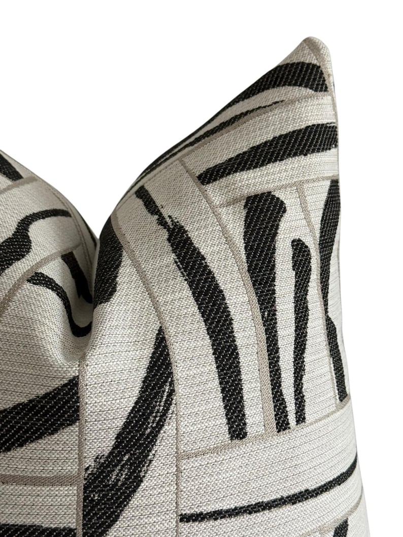 May include: Close-up of a decorative pillow with a geometric pattern. The pillow features a light beige background with bold black brushstroke-style stripes and thin, light gray lines. The pillow has a textured appearance and a soft, inviting look.