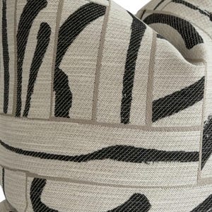 May include: Close-up of a decorative pillow with a cream-colored background and a bold black and gray abstract pattern. The design features thick, curved black brushstrokes and thin gray lines, creating a modern, textured look. The pillow is a square shape.