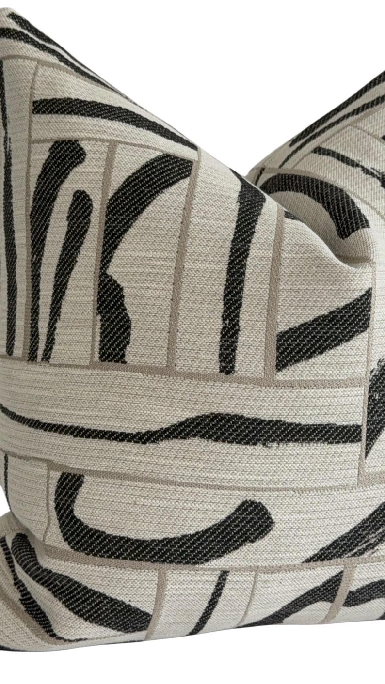 May include: Close-up of a decorative pillow with a cream-colored background and a bold black and gray abstract pattern. The design features thick, curved black brushstrokes and thin gray lines, creating a modern, textured look. The pillow is a square shape.