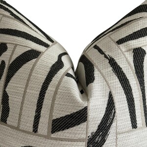 May include: Close-up of a decorative pillow with a geometric pattern. The pillow features a light beige background with bold black and gray brushstroke-style lines. The design creates a modern, abstract aesthetic, suitable for home decor.