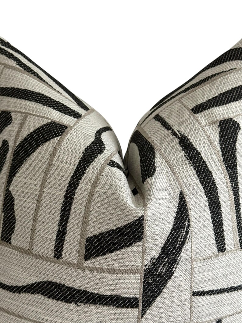May include: Close-up of a decorative pillow with a geometric pattern. The pillow features a light beige background with bold black and gray brushstroke-style lines. The design creates a modern, abstract aesthetic, suitable for home decor.