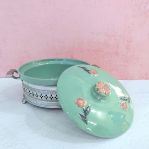 May include: A round, light green ceramic casserole dish with a matching lid featuring small, raised pink floral accents. The dish is set within a silver-toned metal base that has a decorative black and white geometric pattern and two side handles.