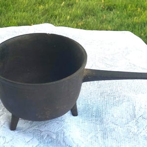 May include: A small, black cast iron pot with a long handle and three short legs. The pot has a rounded body and a wide opening. The handle extends horizontally from the side. The pot is sitting on a white surface.