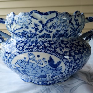 May include: A blue and white ceramic bowl with handles, featuring a scalloped rim and intricate floral patterns. The bowl has a detailed design with a central oval panel depicting a bird and flowers. The overall aesthetic is reminiscent of classic porcelain.