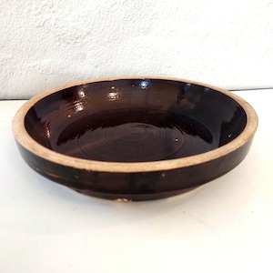 May include: A shallow, circular ceramic bowl featuring a deep, glossy dark brown glaze on the interior and exterior surfaces, with a contrasting unglazed, light beige rim that encircles the top edge of the vessel, creating a distinct border.