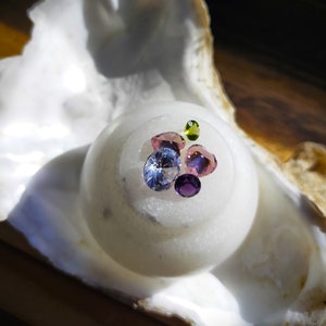 Può includere: Una collezione di pietre preziose colorate, tra cui viola, rosa, blu e verde, è esposta su una superficie rotonda bianca. Le pietre preziose sono disposte a grappolo, con la pietra preziosa più grande al centro.