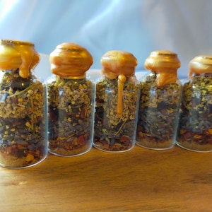 May include: Six small glass bottles with gold lids filled with dried herbs and flowers. The bottles are arranged in a row on a wooden surface.