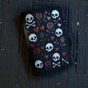Op de afbeelding: Zwarte portemonnee met een herhaald patroon van witte schedels, rode harten en rozen. De portemonnee heeft ook gekruiste botten en een zwarte band. De achtergrond is een donker, getextureerd oppervlak.