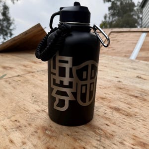 May include: A black insulated water bottle with a silver graphic design. The bottle has a black lid, a black handle, and a black braided strap. A silver carabiner clip is attached to the bottle. The bottle is sitting on a wooden surface.