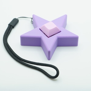 May include: A purple star-shaped keychain with a black lanyard. The star has a small, square, pink keycap in the center.