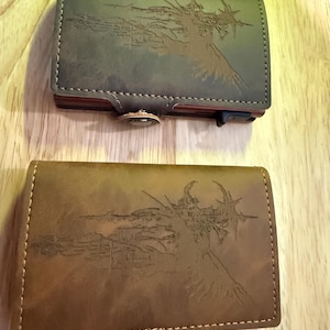 May include: Two leather wallets with a landscape design etched on the front. One wallet is olive green, the other is brown. Both have a button closure and a stitched border. The wallets are on a wooden surface.