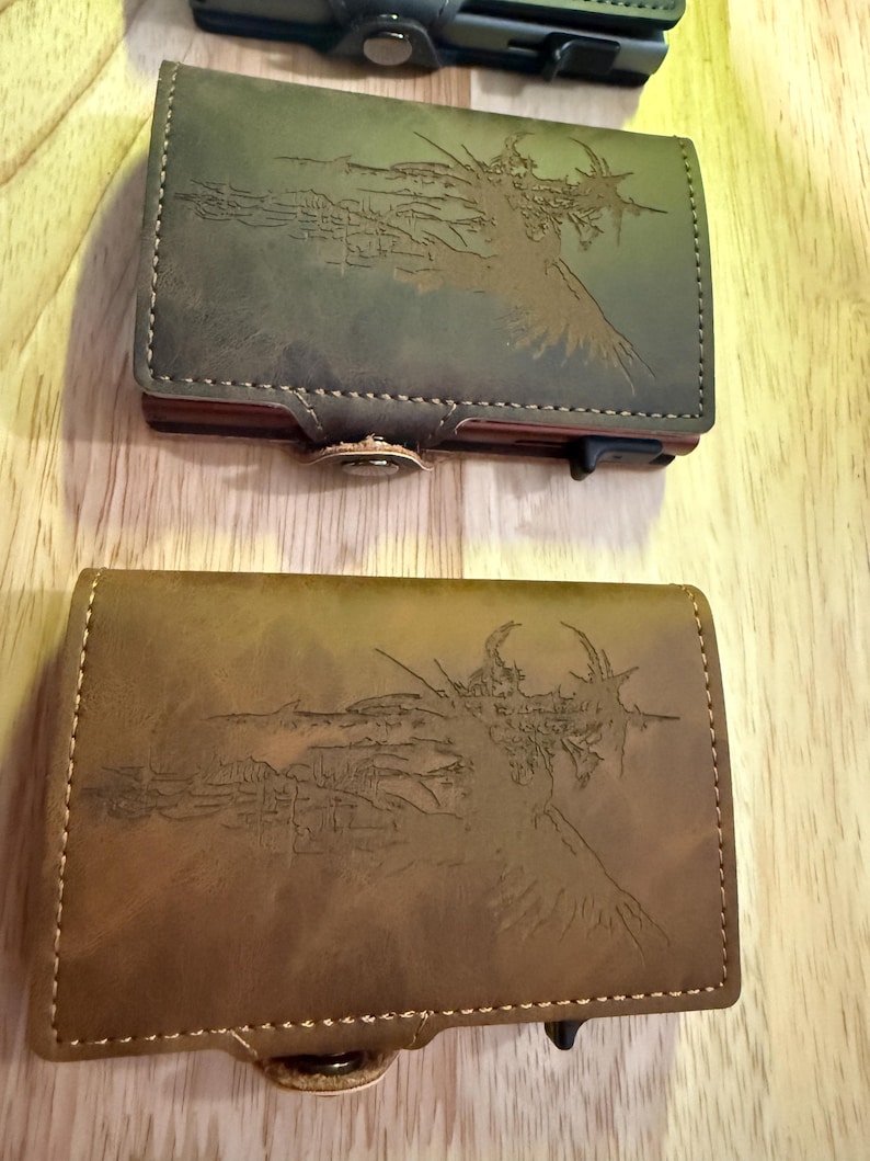 May include: Two leather wallets with a landscape design etched on the front. One wallet is olive green, the other is brown. Both have a button closure and a stitched border. The wallets are on a wooden surface.