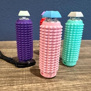May include: Three colorful, textured massage tools. The tools are cylindrical with a spiky surface and a square top. Colors include purple, pink, and teal. A black lanyard is attached to the purple tool. The tools are on a wooden surface.
