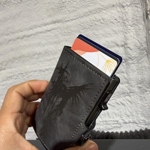 May include: A grey cardholder with a black graphic design. The cardholder is holding two cards with colourful designs. The cardholder has a push button to release the cards.