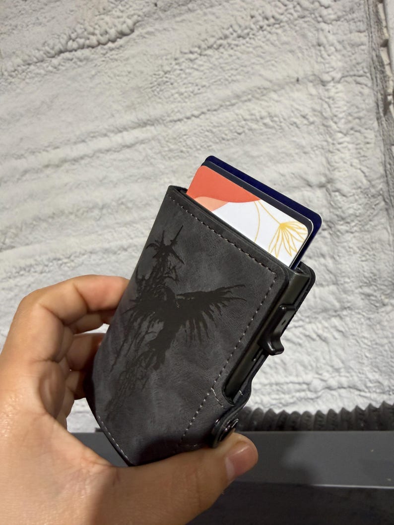 May include: A grey cardholder with a black graphic design. The cardholder is holding two cards with colourful designs. The cardholder has a push button to release the cards.