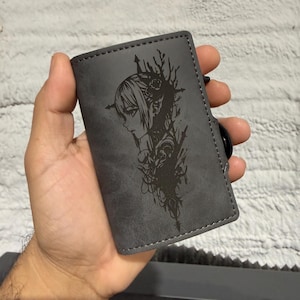 May include: A grey wallet featuring a black etched design of a woman's profile with floral details. The rectangular wallet has a stitched edge and a black button closure. The design is etched into the wallet's surface.