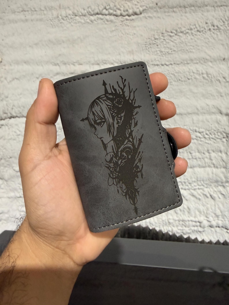May include: A grey wallet featuring a black etched design of a woman's profile with floral details. The rectangular wallet has a stitched edge and a black button closure. The design is etched into the wallet's surface.