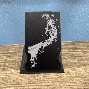 Peut inclure: Support de téléphone en métal noir avec un motif de papillon et de pistolet. Les mots "Memento Mori" sont imprimés sur le support. Le support est posé sur une surface en bois avec un fond bleu.