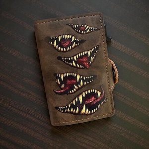 May include: A brown leather wallet decorated with several monster mouth designs, each with sharp, yellow teeth and red tongues. The wallet has a snap closure. The background is dark brown. The design theme is horror or fantasy.