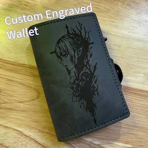 May include: A grey custom engraved wallet featuring a black illustration of a woman's profile and floral details. The text "Custom Engraved Wallet" is in white. The wallet has a black ring attached.