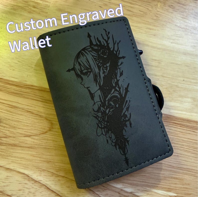 May include: A grey custom engraved wallet featuring a black illustration of a woman's profile and floral details. The text "Custom Engraved Wallet" is in white. The wallet has a black ring attached.
