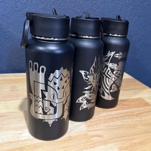 May include: Three black water bottles with silver etched designs. Each bottle has a different design, including a stylized creature and floral patterns. The bottles have black lids and are displayed on a wooden surface.