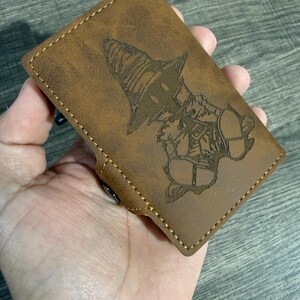 May include: Brown leather wallet featuring a laser-etched character design. The wallet has a stitched border and a snap closure. The design is a detailed illustration of a character with a large hat.