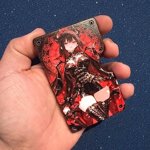 May include: A rectangular, black and red card with a detailed anime-style illustration of a woman in a gothic outfit. The card has a dark red background with a circular design and is held in a hand.