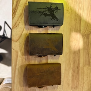 May include: Three leather wallets in green, brown, and tan, each with a black graphic design. The rectangular wallets have clasp closures, displayed against a light wood grain background.