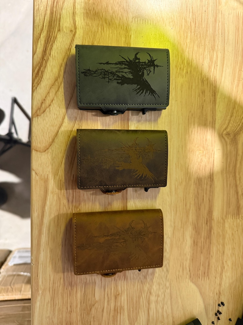 May include: Three leather wallets in green, brown, and tan, each with a black graphic design. The rectangular wallets have clasp closures, displayed against a light wood grain background.