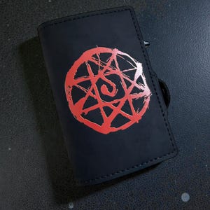 Puede incluir: Cartera negra con un diseño circular rojo. El diseño es un símbolo estilizado con una espiral en el centro. La cartera es rectangular y parece estar hecha de cuero o un material similar.