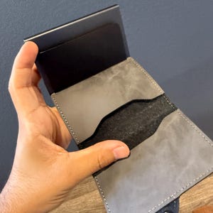 May include: A grey leather wallet with a black metal card holder. The wallet is open, showing a black interior and grey leather pockets. The wallet is held in a hand, highlighting the design and texture of the materials.