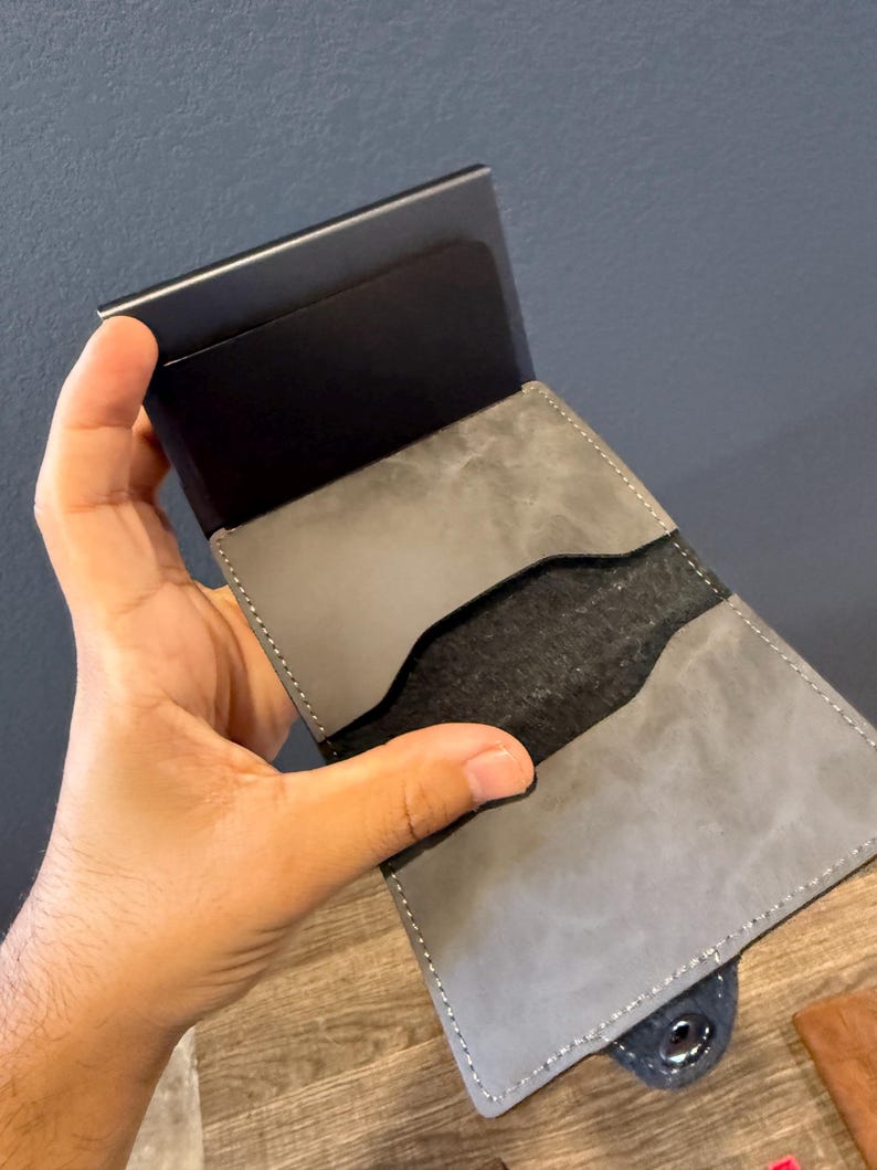 May include: A grey leather wallet with a black metal card holder. The wallet is open, showing a black interior and grey leather pockets. The wallet is held in a hand, highlighting the design and texture of the materials.