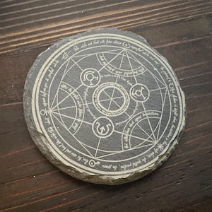 Peut inclure: Un dessous de verre rond en ardoise grise avec un motif circulaire gravé en blanc. Le motif présente un motif géométrique complexe avec des symboles et du texte. Le texte dit "The sole that is but with false allure..."