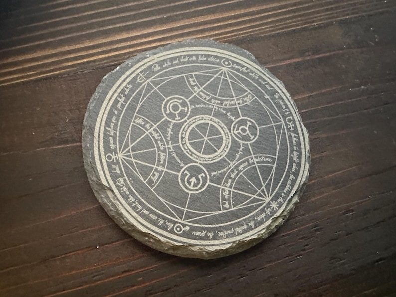 May include: A round, gray slate coaster with a white, engraved, circular design. The design features a complex geometric pattern with symbols and text. The text reads "The sole that is but with false allure..."