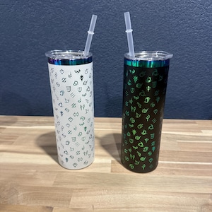 Könnte beinhalten: Zwei Edelstahlbecher mit einem grün-weißen Muster aus Symbolen. Ein Becher ist weiß mit grünen Symbolen und der andere ist schwarz mit grünen Symbolen. Beide Becher haben durchsichtige Plastikstrohhalme.