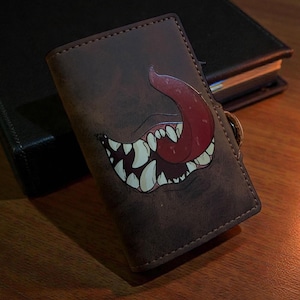 May include: Brown leather wallet featuring a cartoon monster mouth design with sharp white teeth and a red tongue. The wallet is rectangular with a stitched border and a small loop. The background includes a dark book on a wooden surface.