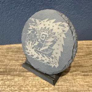 May include: A gray, oval-shaped slate coaster with a detailed, etched design of a creature. The coaster is displayed on a small, dark gray stand, showcasing the artwork. The design is a light color, contrasting against the darker slate.