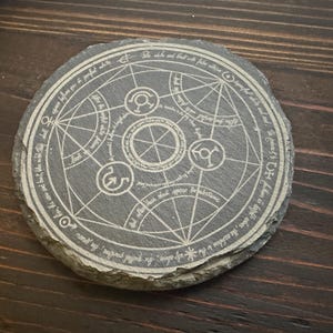 May include: A round, gray slate coaster with a white, engraved alchemical symbol. The symbol is surrounded by a circle of text in white.