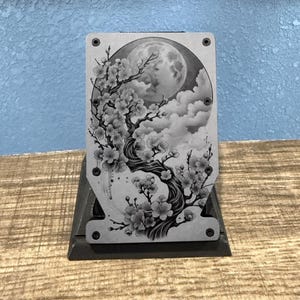 May include: A metal card holder with a black and white cherry blossom design. The design features a full moon, clouds, and a blossoming tree. The card holder is mounted on a dark gray stand.