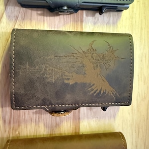 May include: A selection of leather wallets in green and brown hues. The middle wallet showcases a detailed brown graphic design. Each wallet has a button closure and a stitched border. The wallets are displayed on a wooden surface.