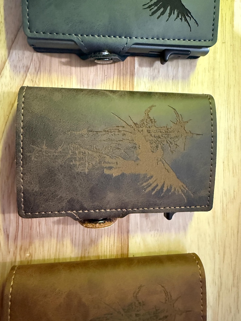 May include: A selection of leather wallets in green and brown hues. The middle wallet showcases a detailed brown graphic design. Each wallet has a button closure and a stitched border. The wallets are displayed on a wooden surface.