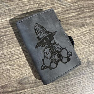 Op de afbeelding: Grijze portemonnee met een zwarte illustratie van een personage uit Final Fantasy. De rechthoekige portemonnee heeft een gestikte rand. Het personage draagt een hoed en gewaden.